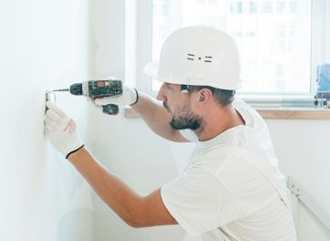 An engineer in a white hard hat and gloves using a nail gun to fasten a fixture onto a white wall indoors.